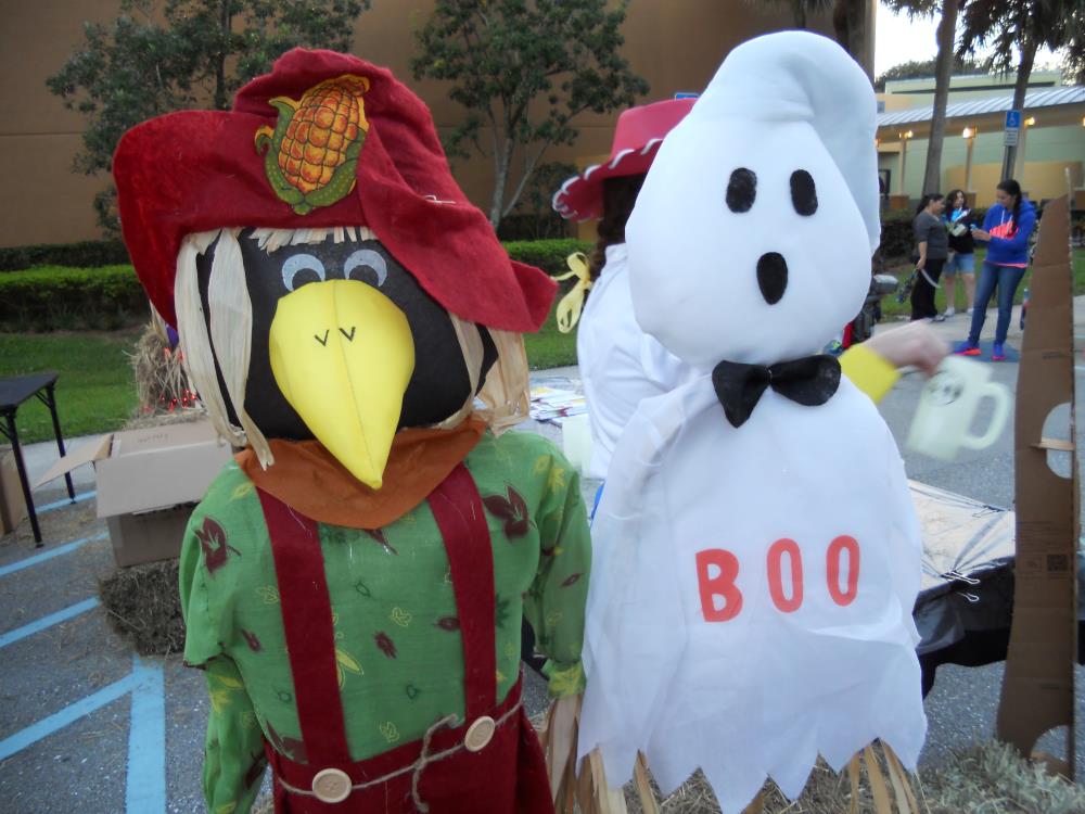 Crow dressed in scarecrow lawn decoration and white ghost with black bowtie lawn decoration with text 